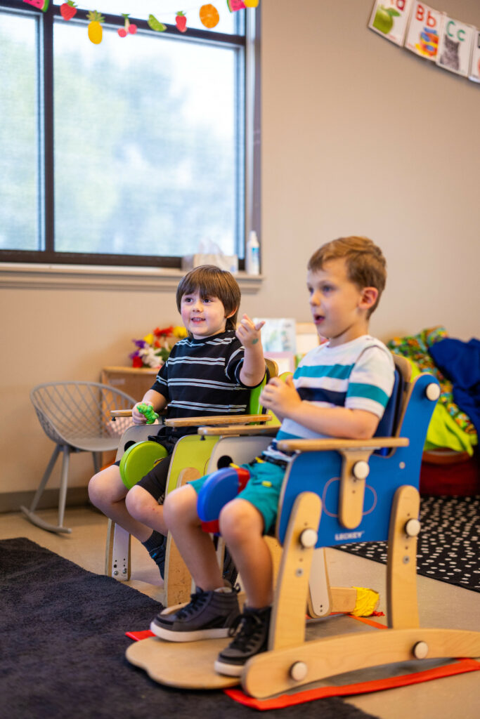 Two young boys sitting in adjustable chairs with a foot rest, arm rests, and pommels.